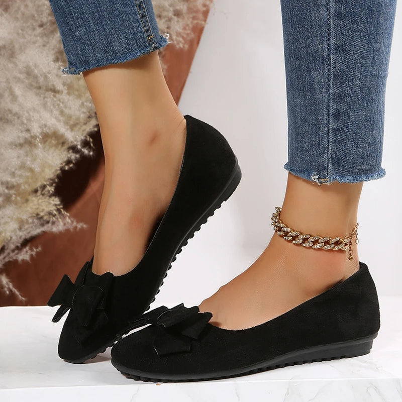 Chaussures ballerines noires en daim avec nœud, portées avec un jean bleu effiloché et bracelet doré, mode féminine élégante et décontractée.