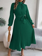 Robe longue verte plissée avec manches longues, ceinture à nouer, portée par une femme tenant un sac en osier, intérieur moderne.