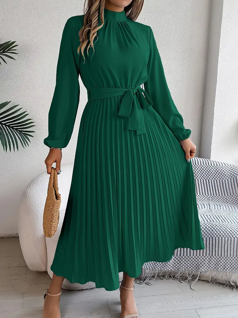 Robe longue verte plissée avec manches longues, ceinture à nouer, portée par une femme tenant un sac en osier, intérieur moderne.