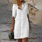 Robe chemise blanche en lin portée par une femme sur un chemin pavé, style décontracté, manches retroussées, accessoirisée de bracelets et chapeau.