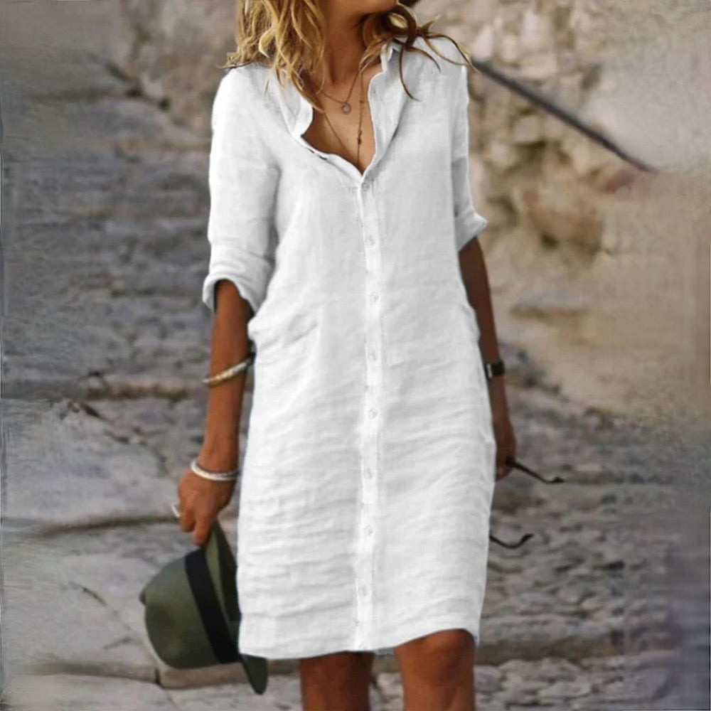 Robe chemise blanche en lin portée par une femme sur un chemin pavé, style décontracté, manches retroussées, accessoirisée de bracelets et chapeau.