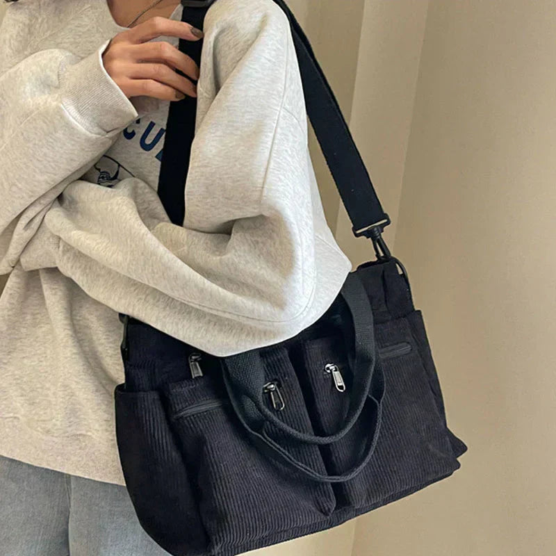 Sac à bandoulière noir en velours côtelé porté par une personne en sweat gris. Mode décontractée, accessoires tendance, style urbain.