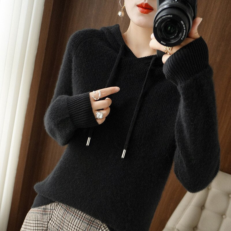 Femme en pull noir à capuche, tenant un appareil photo, portant des bagues argentées. Mode automne, style décontracté, intérieur moderne.