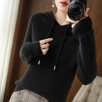 Femme en pull noir à capuche, tenant un appareil photo, portant des bagues argentées. Mode automne, style décontracté, intérieur moderne.