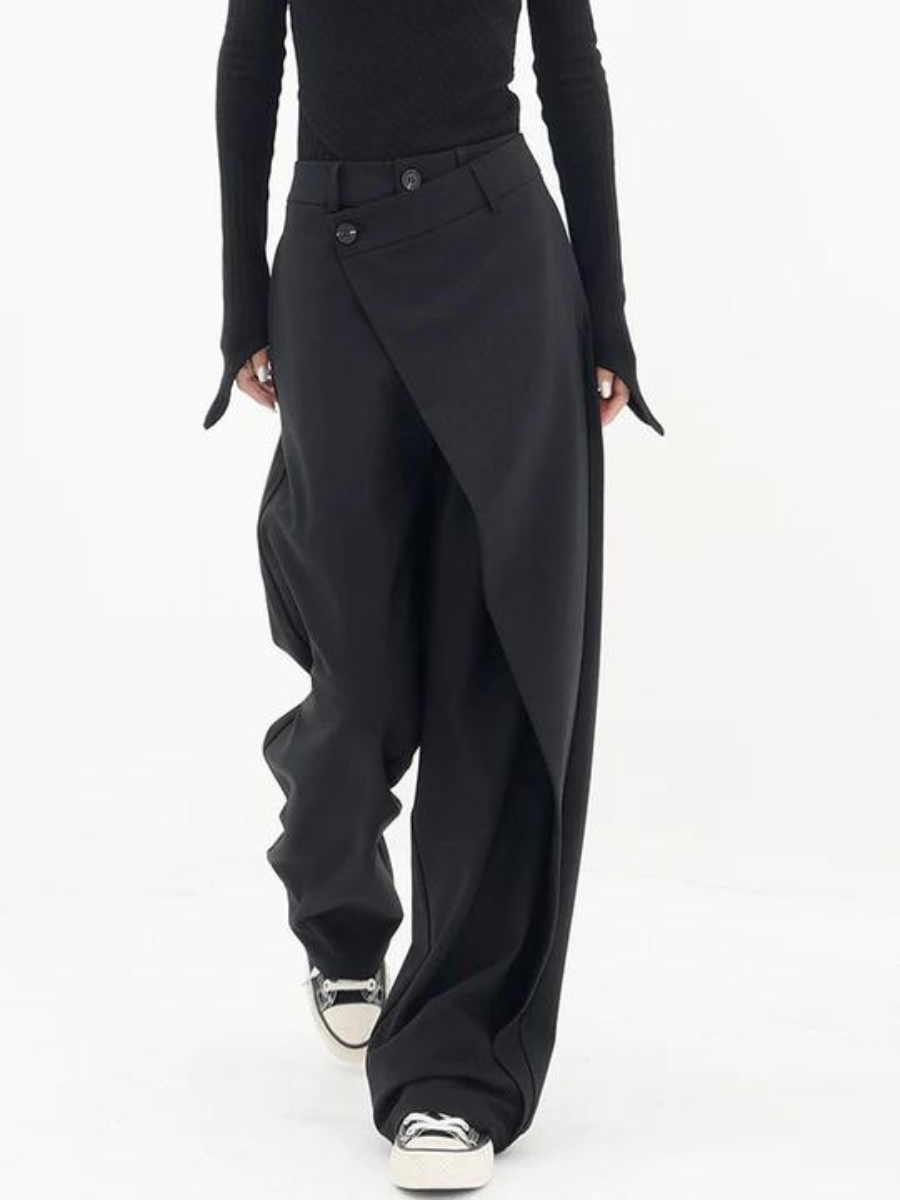 Pantalon noir ample et asymétrique pour femme, style moderne et élégant, avec boutons décoratifs, porté avec un haut noir et des baskets blanches.