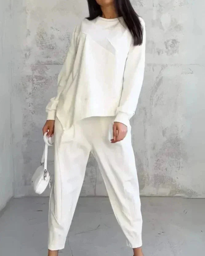 Femme en tenue décontractée blanche, portant un pull ample et un pantalon, avec un petit sac à main blanc, posant devant un mur texturé gris.
