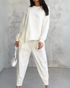 Femme en tenue décontractée blanche, portant un pull ample et un pantalon, avec un petit sac à main blanc, posant devant un mur texturé gris.