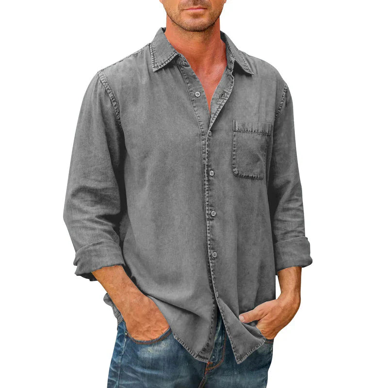 Homme portant une chemise en jean gris délavé avec manches longues retroussées, poche poitrine, et jeans bleus. Mode décontractée masculine.