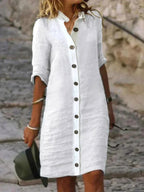 Robe chemise blanche en lin avec boutons, manches retroussées, portée par une femme sur un chemin pavé. Mode estivale élégante et décontractée.