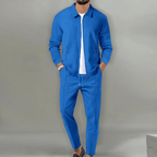 Homme portant un costume bleu moderne avec veste et pantalon assortis, sur fond gris. Mode masculine élégante, tenue décontractée chic.