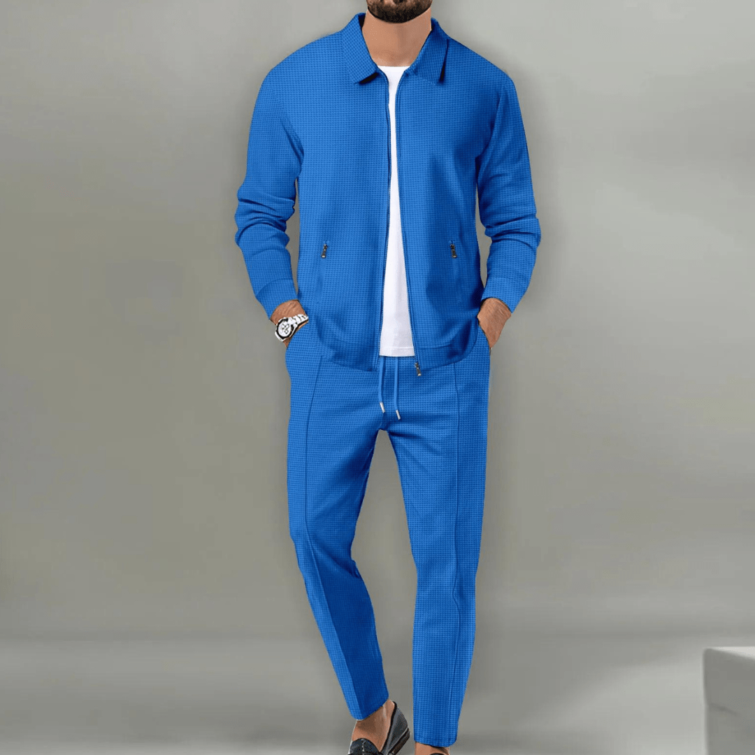 Homme portant un costume bleu moderne avec veste et pantalon assortis, sur fond gris. Mode masculine élégante, tenue décontractée chic.