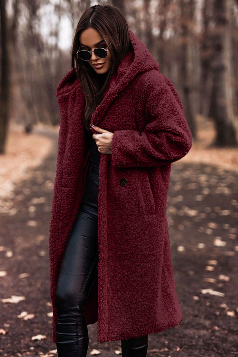 Femme portant un manteau en fausse fourrure bordeaux, lunettes de soleil, pantalon noir, marchant dans un parc automnal. Mode hiver élégante.