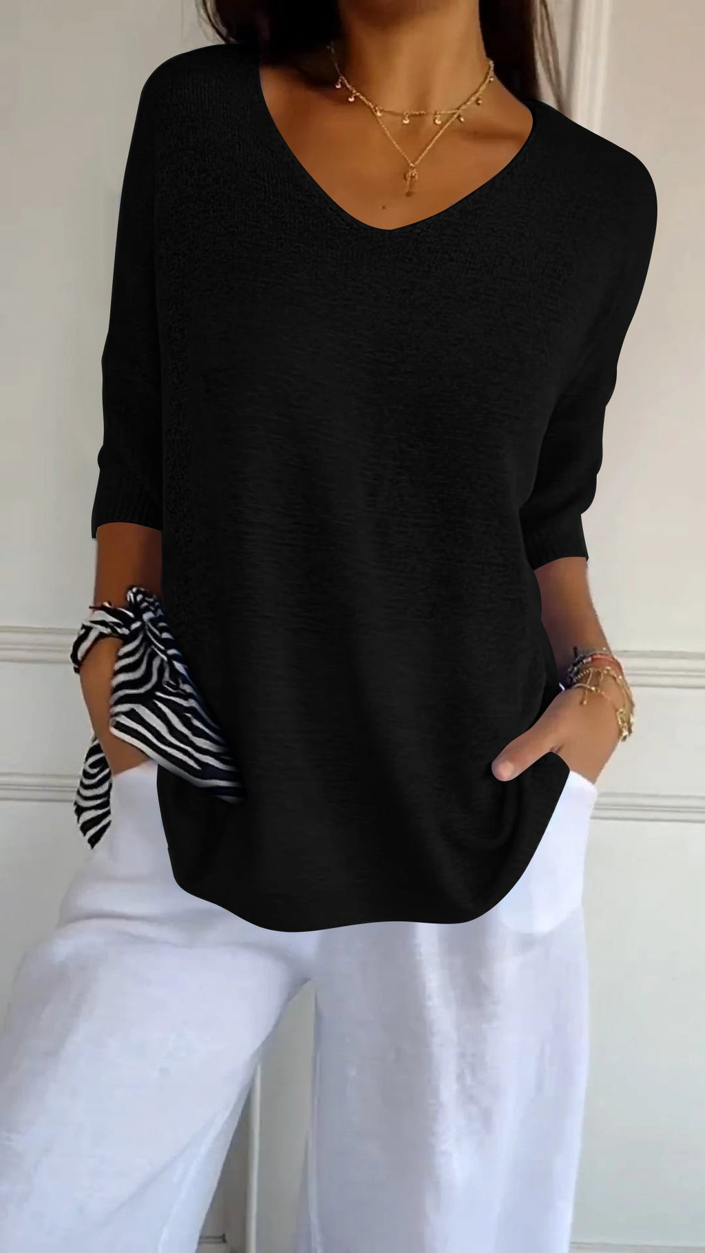 Femme portant un pull noir à col en V, manches retroussées, avec un pantalon blanc. Style décontracté, chic, mode féminine tendance.