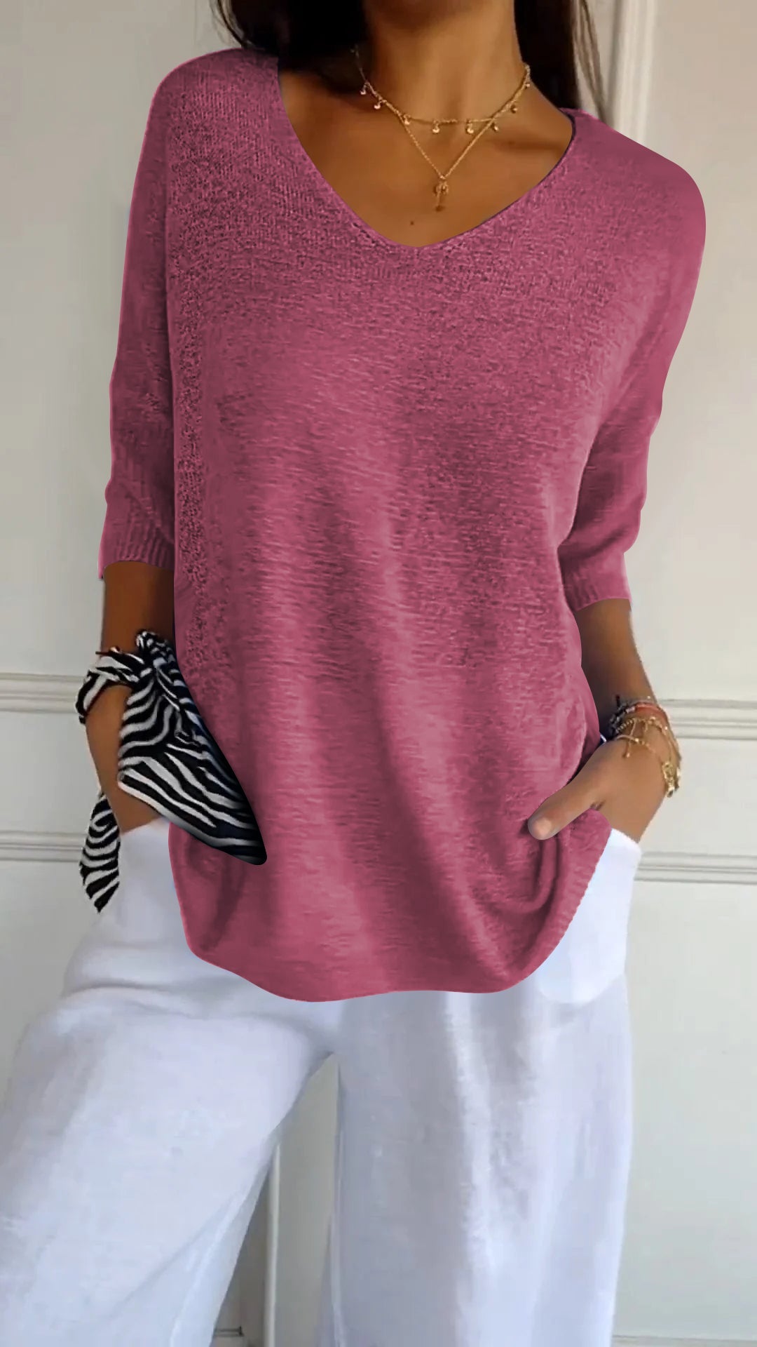 Femme portant un pull rose à manches longues avec un col en V, tenant un foulard rayé noir et blanc, pantalon blanc, style décontracté chic.