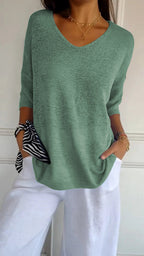 Femme portant un pull vert à col en V, manches trois-quarts, avec un pantalon blanc. Accessoire: foulard rayé noir et blanc au poignet.