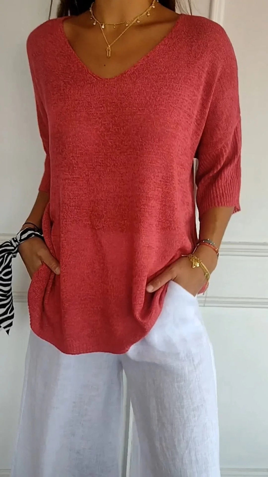 Femme portant un pull rouge à manches trois-quarts et un pantalon blanc en lin, accessoirisé d'un bracelet et d'un foulard zébré au poignet.