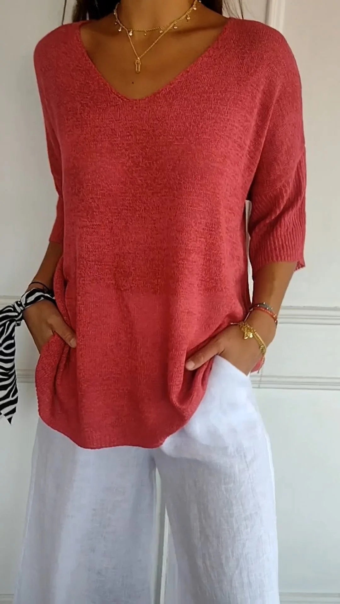 Femme portant un pull rouge à manches trois-quarts et un pantalon blanc en lin, accessoirisé d'un bracelet et d'un foulard zébré au poignet.