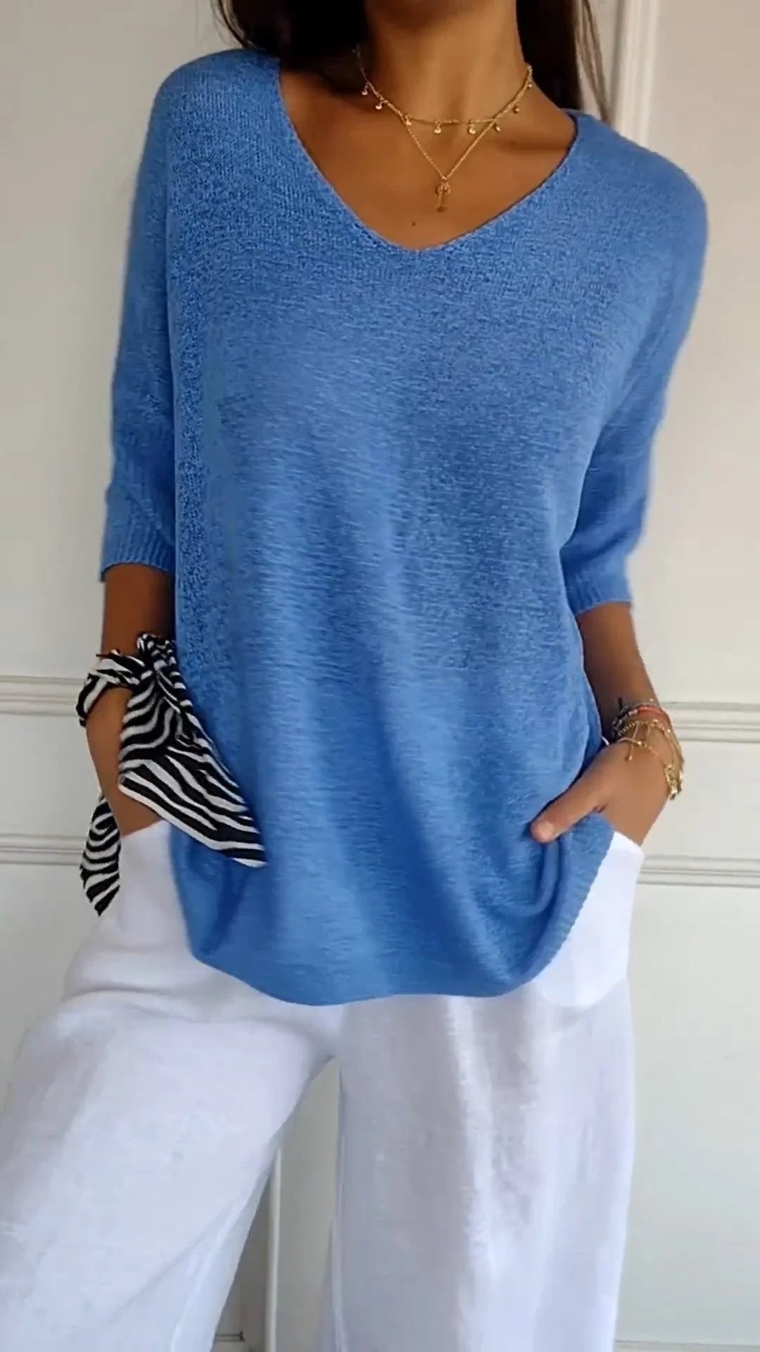 Femme portant un pull bleu ample avec manches trois-quarts, pantalon blanc, et foulard rayé noir et blanc au poignet. Mode décontractée élégante.