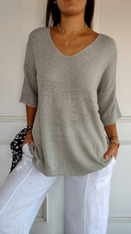 Femme portant un pull gris à manches trois-quarts et un pantalon blanc, accessoirisée de colliers dorés et bracelets colorés. Mode décontractée élégante.