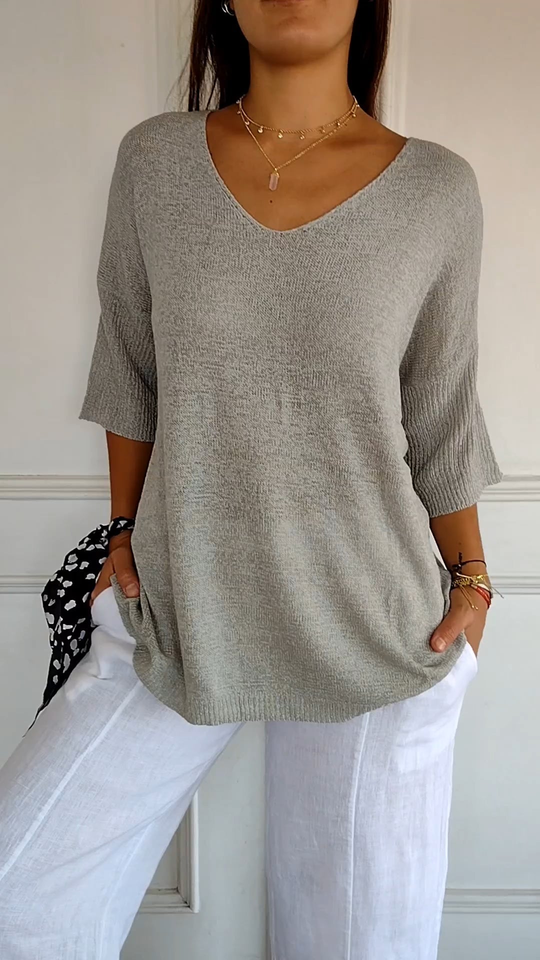 Femme portant un pull gris à manches trois-quarts et un pantalon blanc, accessoirisée de colliers dorés et bracelets colorés. Mode décontractée élégante.