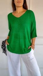 Femme portant un pull vert à col en V et manches trois-quarts, associé à un pantalon blanc. Mode décontractée, tendance printemps-été.