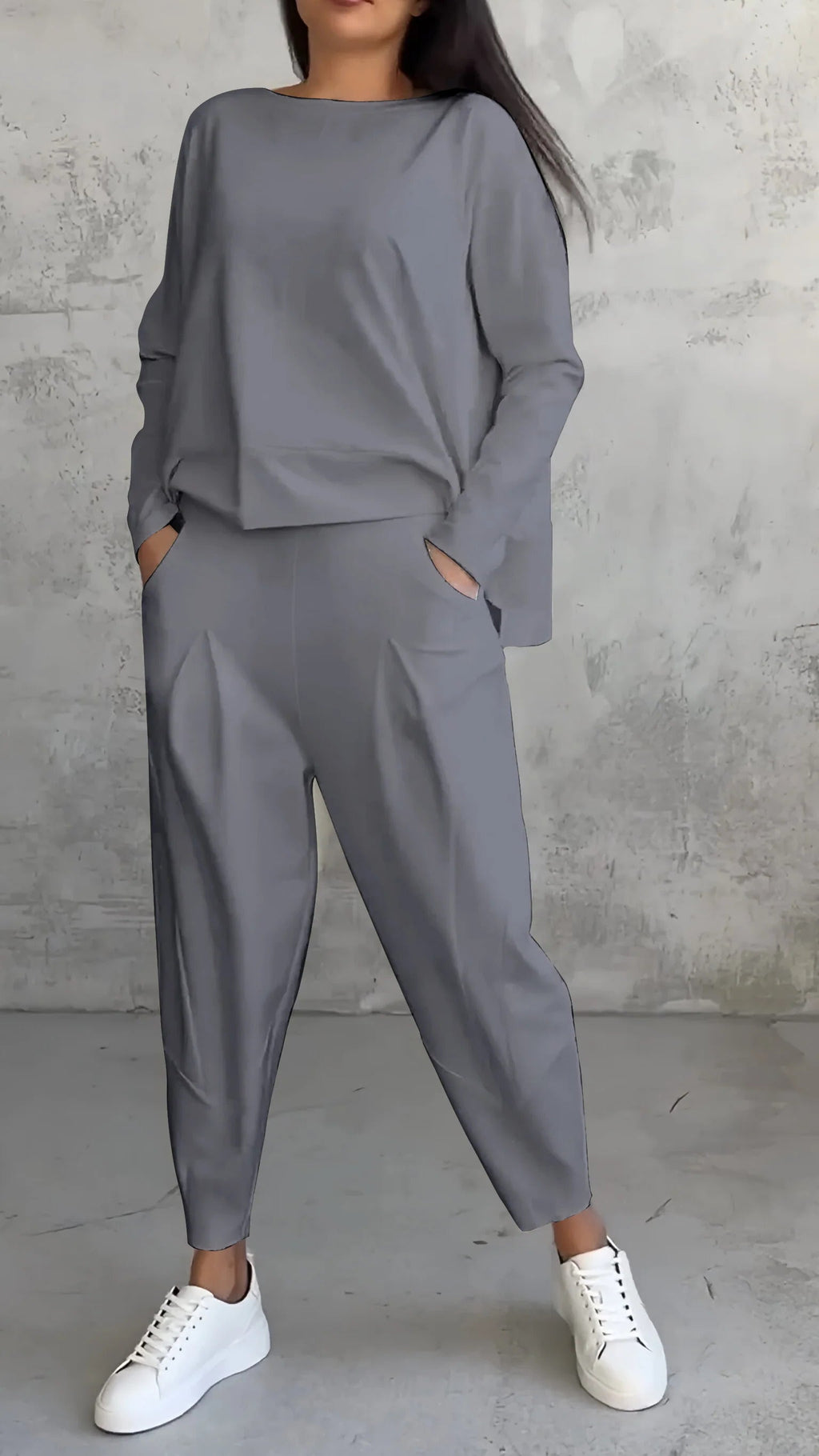 Femme portant un ensemble gris élégant avec un haut ample et un pantalon à pinces, posant contre un mur texturé, chaussures blanches.