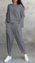 Femme portant un ensemble gris élégant avec un haut ample et un pantalon à pinces, posant contre un mur texturé, chaussures blanches.