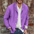 Homme souriant portant une veste en lin violet sur une chemise blanche, posant devant un mur en briques. Mode décontractée, style estival.
