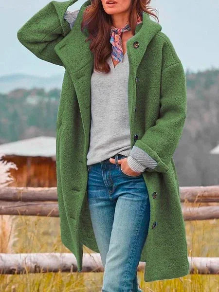 Femme portant un manteau vert en laine, pull gris, jean bleu, foulard coloré, posant en extérieur. Mode automne-hiver, style décontracté.