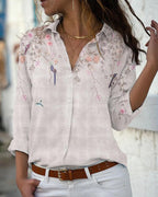 Femme portant une chemise blanche à motifs floraux et oiseaux, manches retroussées, col ouvert, ceinture marron; mode élégante et décontractée.