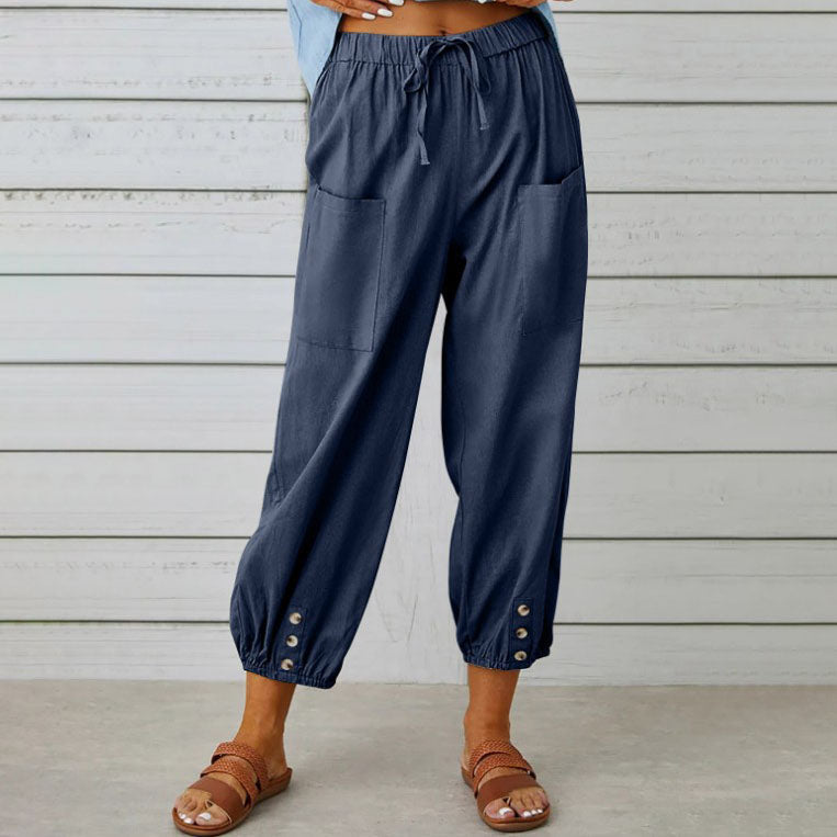 Pantalon ample bleu marine pour femme avec poches avant, boutons décoratifs en bas, porté avec des sandales marron sur fond de mur en bois blanc.