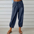 Pantalon ample bleu marine pour femme avec poches avant, boutons décoratifs en bas, porté avec des sandales marron sur fond de mur en bois blanc.
