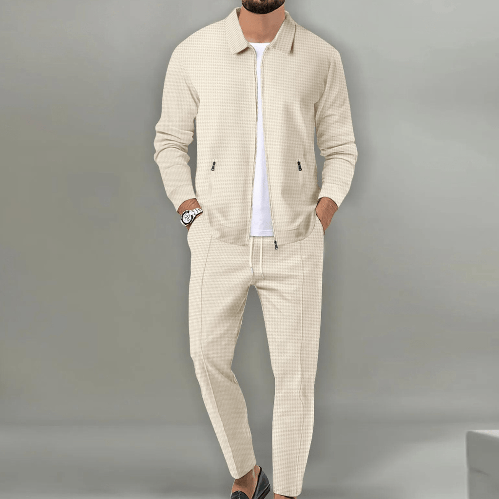 Homme portant un ensemble beige élégant, veste et pantalon assortis, style décontracté chic, montre au poignet, fond gris minimaliste.