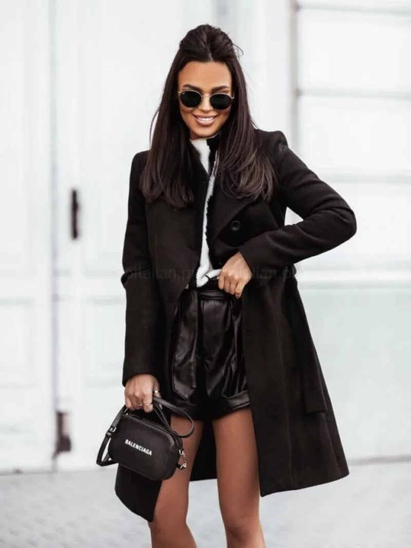 Femme élégante en manteau noir, lunettes de soleil, short en cuir, tenant un sac Balenciaga. Mode chic, style urbain, tendance automne-hiver.