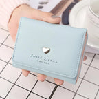 Portefeuille bleu pastel en cuir synthétique avec un motif de cœur doré, tenu par une main sur une table à carreaux. Accessoire mode élégant.
