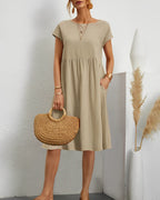 Robe beige décontractée avec manches courtes, portée par une femme tenant un sac en osier. Décor minimaliste avec vase et pampas. Mode été chic.