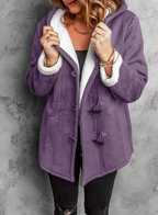 Femme portant un manteau violet en laine avec doublure blanche, boutons en bois, sur fond de mur gris. Mode hiver, style élégant et confortable.