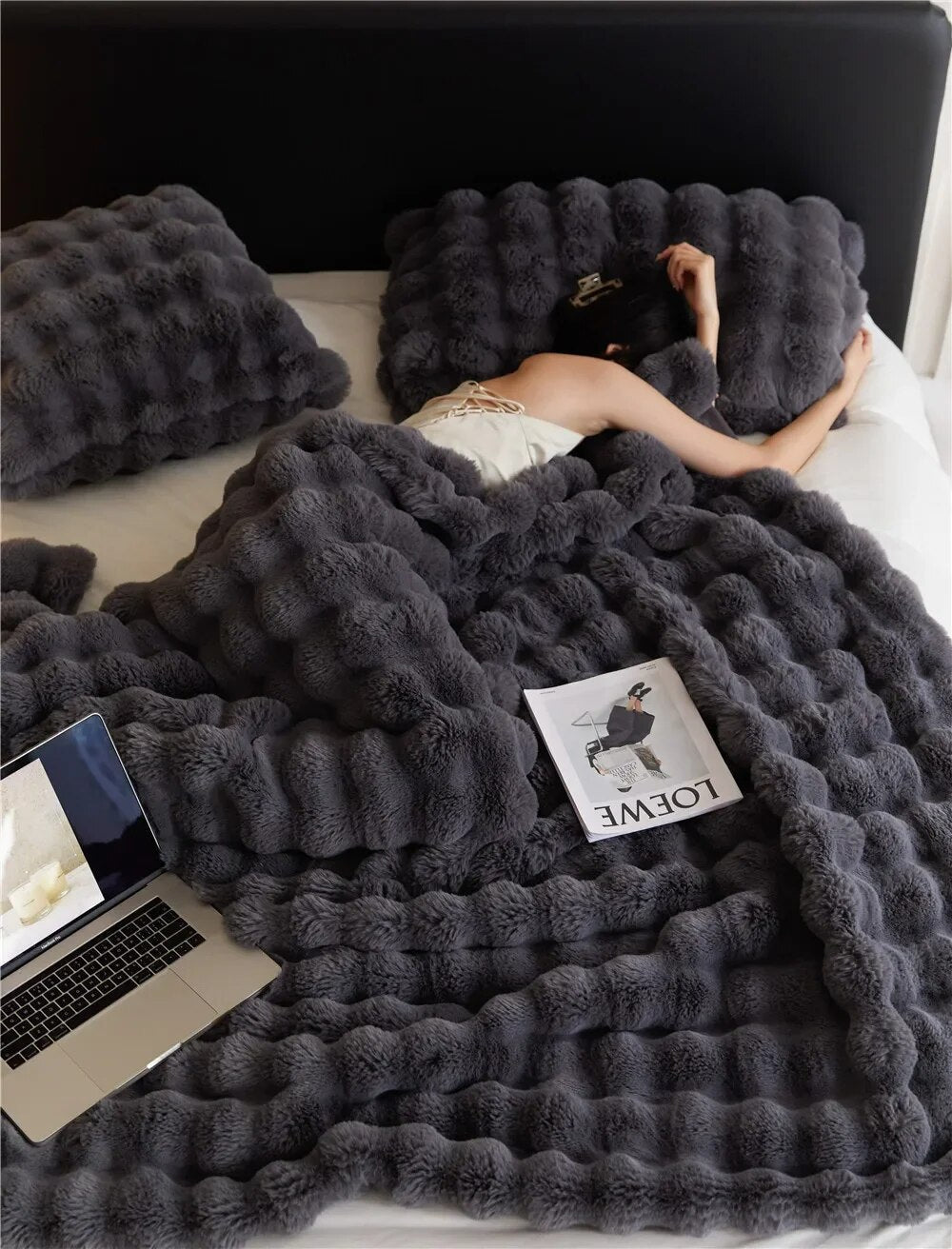 Femme dormant sous une couverture en fausse fourrure grise avec oreillers assortis, magazine et ordinateur portable sur le lit. Confort et détente.