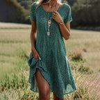 Robe d'été verte à pois blancs portée par une femme dans un champ, accessoirisée avec des colliers et une montre. Mode féminine, style champêtre.