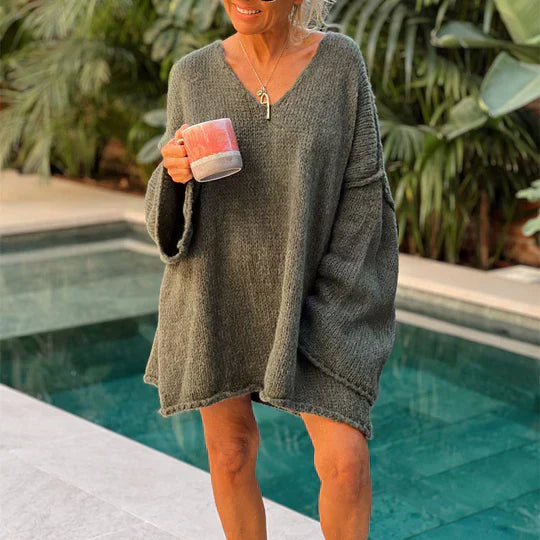 Femme souriante en pull oversize gris, tenant une tasse, debout près d'une piscine entourée de végétation tropicale. Mode détente, style décontracté.