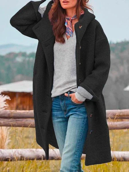 Femme portant un manteau noir élégant, pull gris, jean bleu, foulard coloré, posant devant un paysage champêtre. Mode automne-hiver tendance.