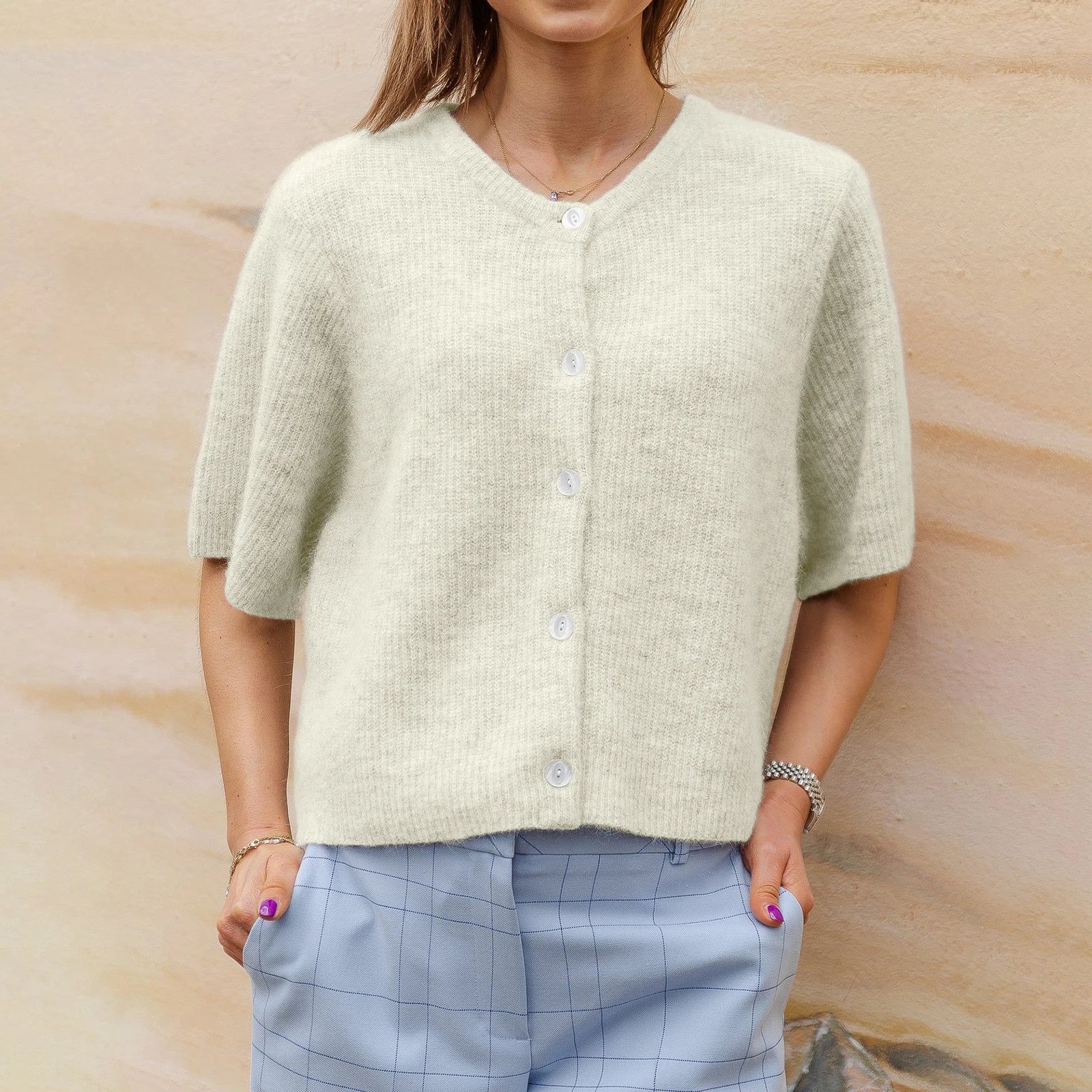 Femme portant un cardigan en laine beige avec boutons, manches courtes, associé à un pantalon bleu à carreaux. Mode automne, tenue décontractée élégante.