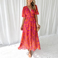 Robe longue d'été rose et orange à motifs floraux, manches courtes bouffantes, décolleté en V, portée par une femme souriante, fond rideaux blancs.