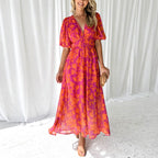 Robe longue d'été rose et orange à motifs floraux, manches courtes bouffantes, décolleté en V, portée par une femme souriante, fond rideaux blancs.