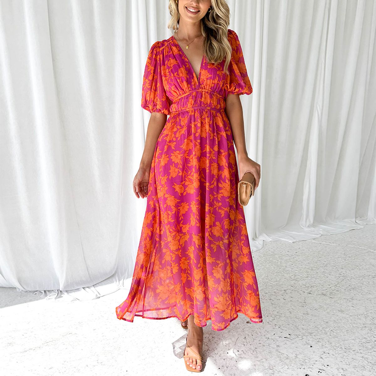 Robe longue d'été rose et orange à motifs floraux, manches courtes bouffantes, décolleté en V, portée par une femme souriante, fond rideaux blancs.