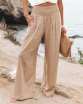 Pantalon large beige en lin pour femme, taille haute, sur une plage. Mode estivale, tenue décontractée, accessoire sac en paille.