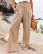 Pantalon large beige en lin pour femme, taille haute, sur une plage. Mode estivale, tenue décontractée, accessoire sac en paille.