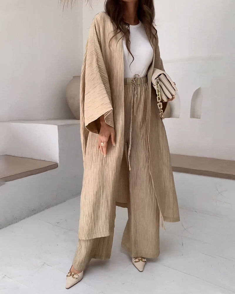 Femme portant un ensemble beige plissé, composé d'un kimono et d'un pantalon ample, avec un sac à main blanc et des talons pointus. Mode élégante et minimaliste.