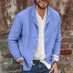 Homme souriant portant une veste en lin bleu sur une chemise blanche, devant un mur en briques. Mode décontractée, style estival, tenue élégante.