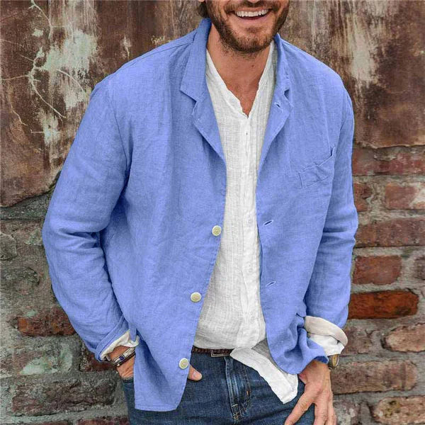 Homme souriant portant une veste en lin bleu sur une chemise blanche, devant un mur en briques. Mode décontractée, style estival, tenue élégante.